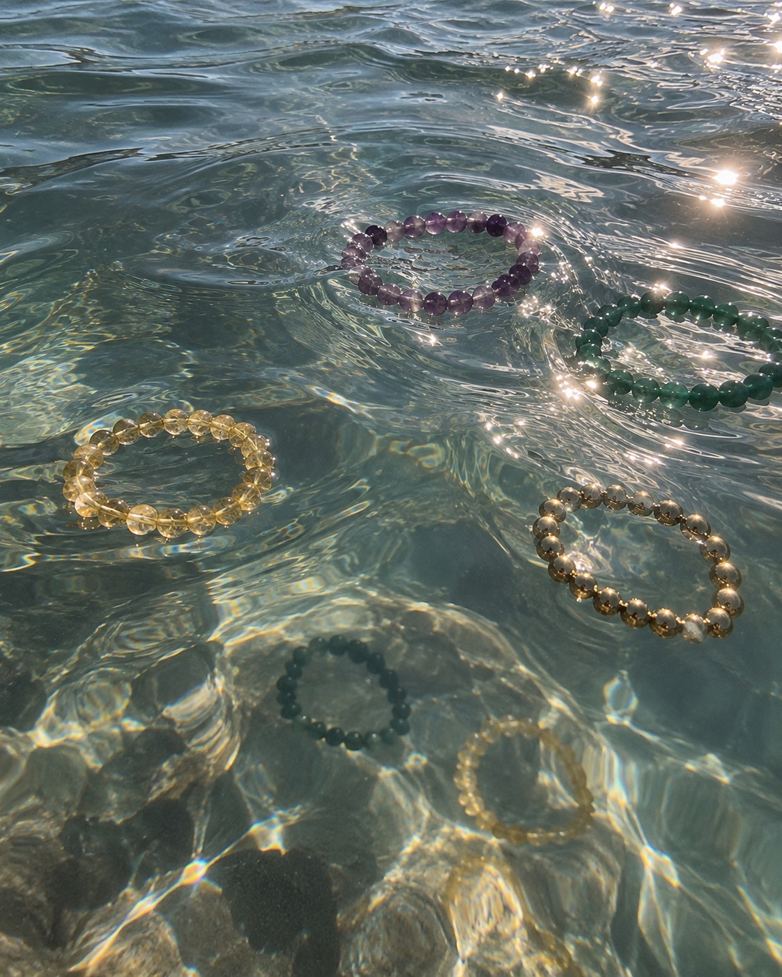 Five beaded gemstone bracelets floating on clear, sunlit sea water